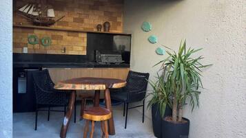 An outdoor patio with a wooden table and chairs, a potted plant, and a brick wall with a grill area.