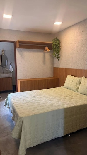 A bedroom with a bed, a wooden headboard, a hanging plant, and a bathroom with a mirror and towel rack. - Casa 7 Villa Natur (São Miguel dos Milagres)