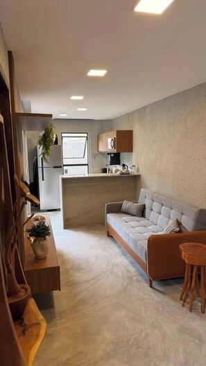 A modern living room with a sofa, wooden side table, and a small coffee table. A kitchen area is visible in the background with a refrigerator and a microwave. - Casa 7 Villa Natur (São Miguel dos Milagres)