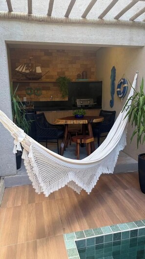A hammock over a wooden table and chairs, a kitchen area with a grill, and a tiled floor. - Casa 7 Villa Natur (São Miguel dos Milagres)