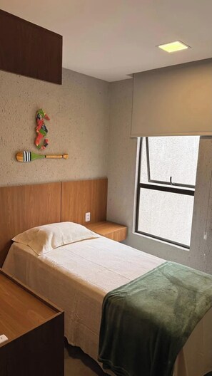Un lit simple avec une tête de lit en bois, une table de chevet, un plaid vert, une fenêtre avec des stores et une pagaie en bois fixée au mur.