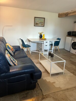 A living room with a black leather sofa, a white table, and a blue chair. There's a painting on the wall and a washing machine in the corner.