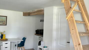Kompakti oleskelutila, jossa on valkoiset seinät, puinen katto, ruokailualue pöytineen ja tuoleineen, kodinkoneilla varustettu keittiö ja puinen tikapuu.