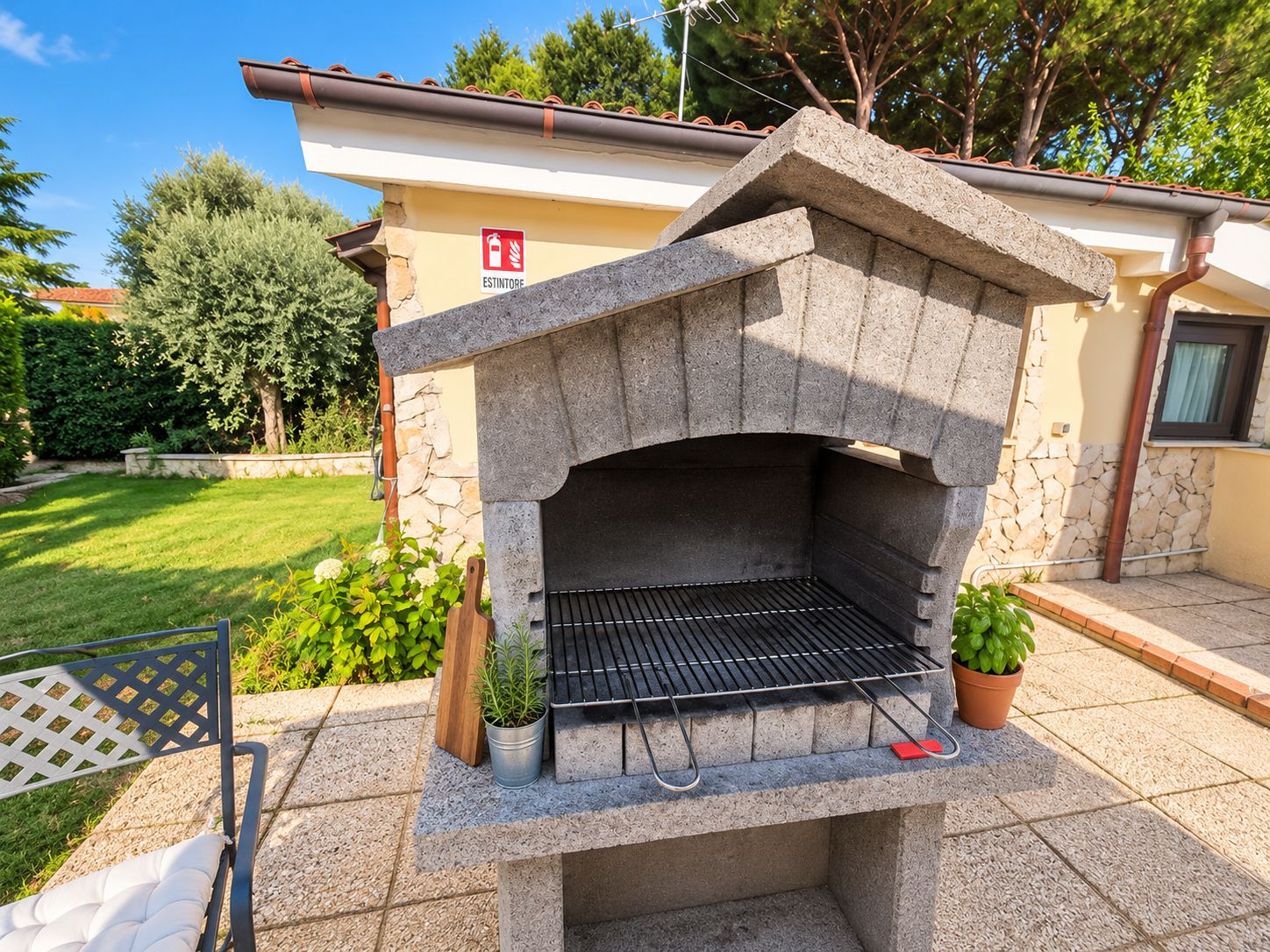 An outdoor stone-built barbecue grill with a metal grate, surrounded by a paved patio area with potted plants and a wooden chair.