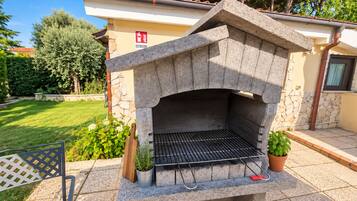 An outdoor stone-built barbecue grill with a metal grate, surrounded by a paved patio area with potted plants and a wooden chair.