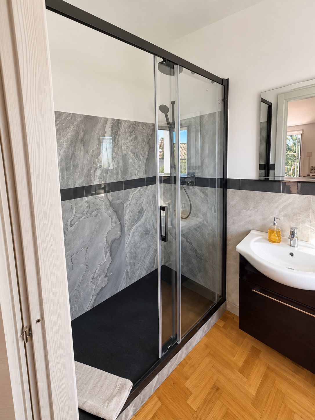 A modern bathroom with a glass-enclosed shower, marble walls, a wooden floor, and a dark-toned vanity with a sink and soap dispenser.