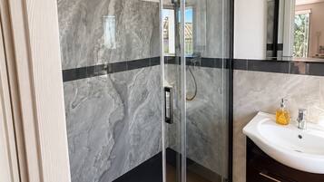 A modern bathroom with a glass-enclosed shower, marble walls, a wooden floor, and a dark-toned vanity with a sink and soap dispenser.