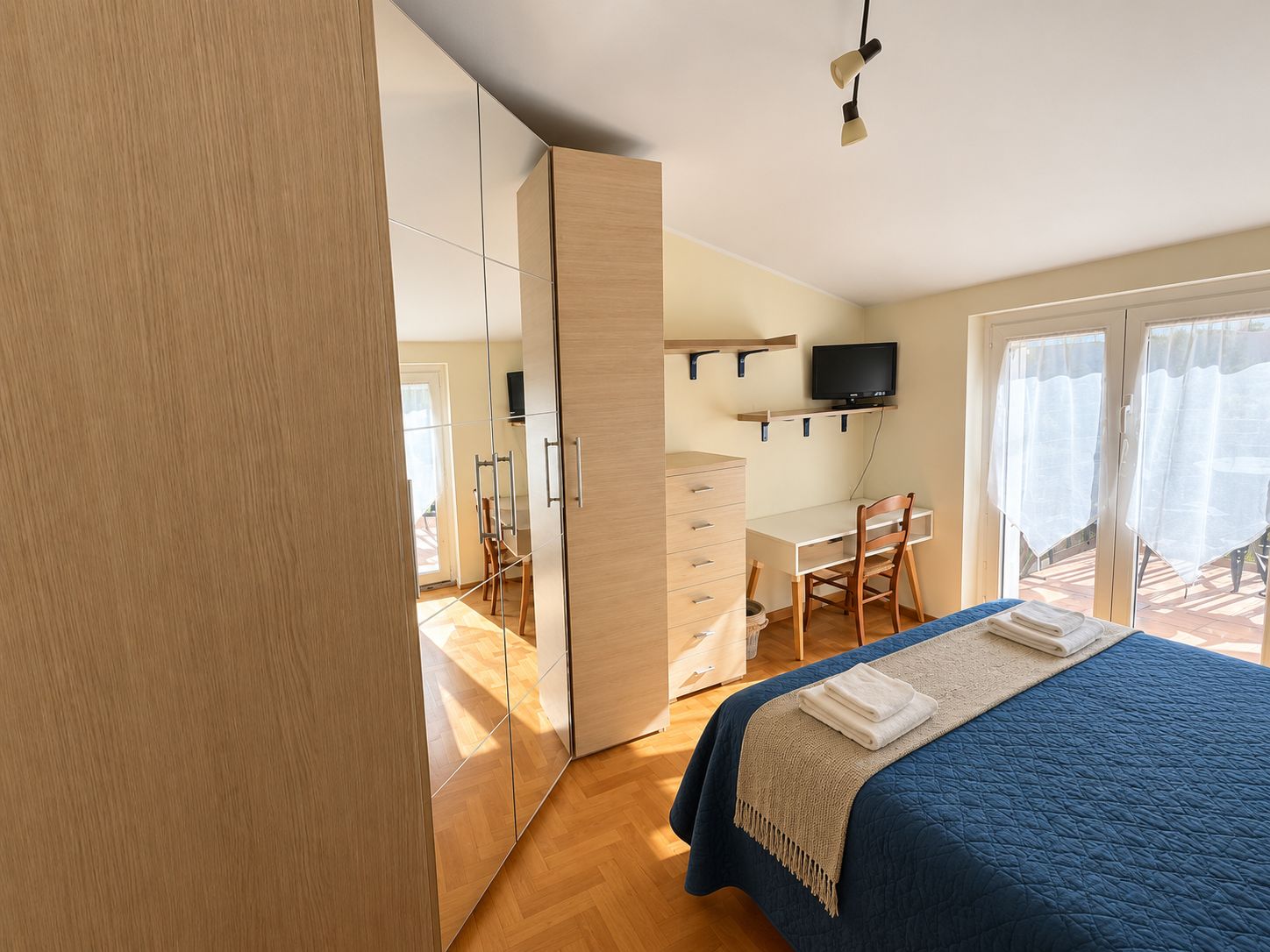 A bedroom with a bed, a desk, a chair, a television, and a sliding door leading to a balcony.