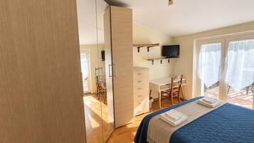 A bedroom with a bed, a desk, a chair, a television, and a sliding door leading to a balcony.