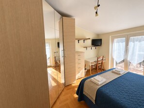 A bedroom with a bed, a desk, a chair, a television, and a sliding door leading to a balcony.