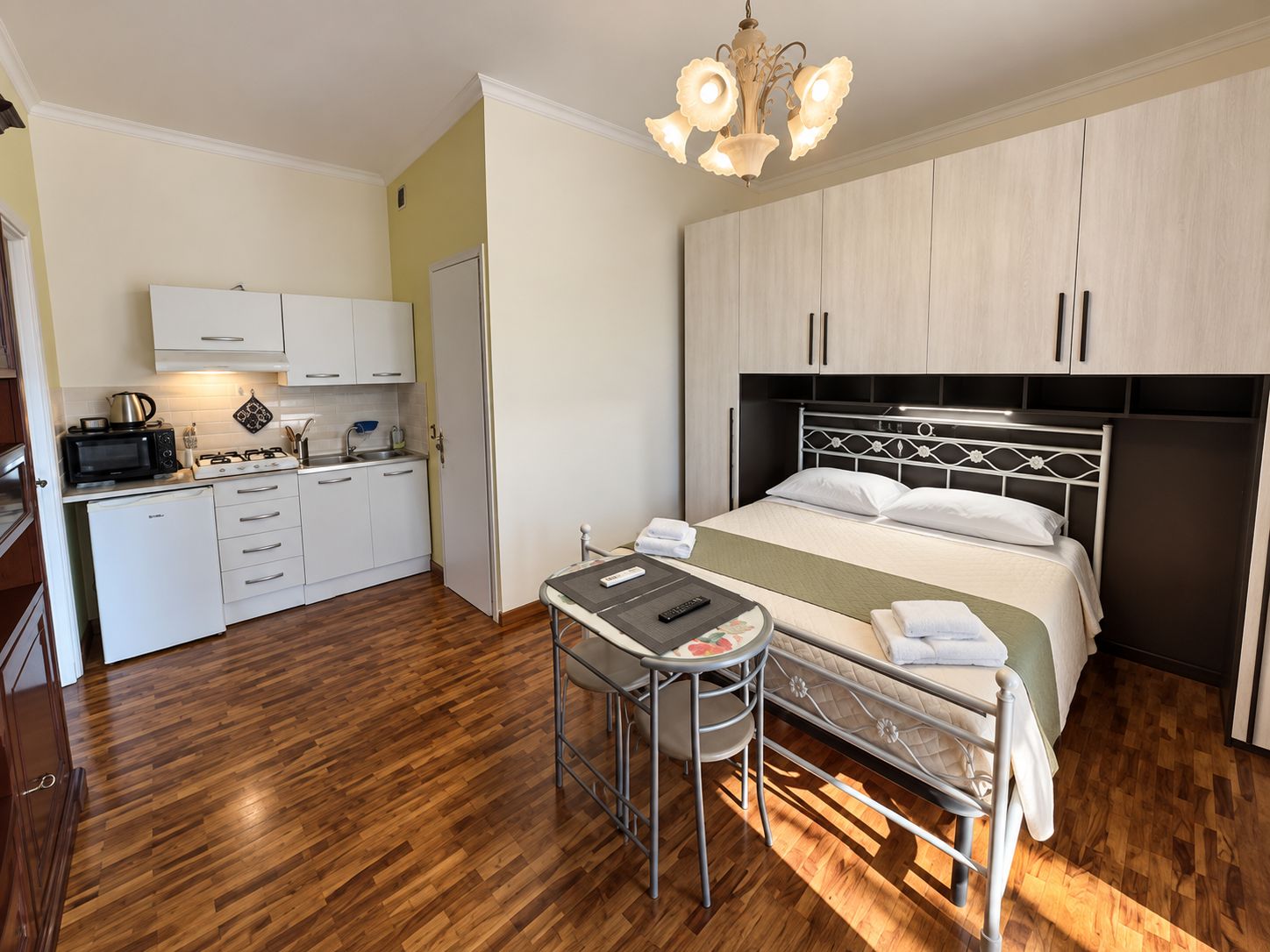 Ein kleines Hotelzimmer mit einer Küchenzeile, einem Bett mit Kopfteil, einem kleinen Tisch mit Stuhl und einer Deckenleuchte.
