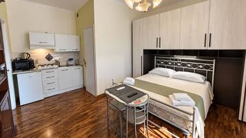 Ein kleines Hotelzimmer mit einer Küchenzeile, einem Bett mit Kopfteil, einem kleinen Tisch mit Stuhl und einer Deckenleuchte.