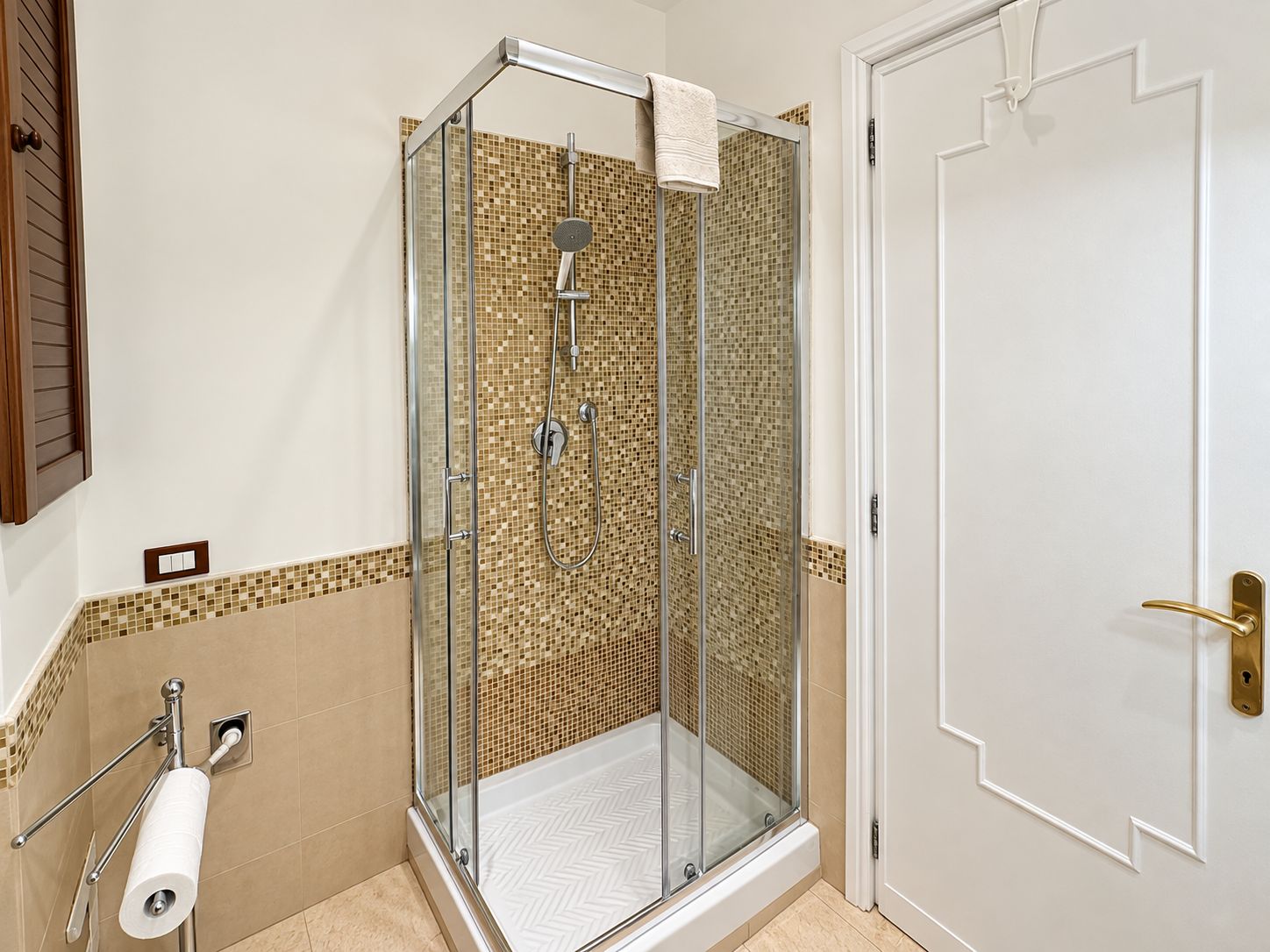 A glass-enclosed shower with a tiled mosaic wall, a towel hanging on the door, and a toilet paper holder on the wall.