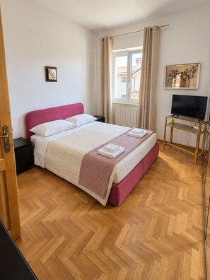 Una habitación de hotel con cama, televisor sobre un soporte, ventana con cortinas y un cuadro en la pared.