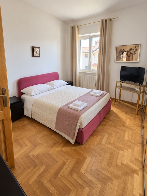 A hotel room with a bed, a television on a stand, a window with curtains, and a painting on the wall.