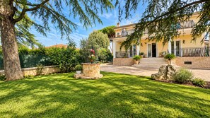 A well-maintained garden with a stone fountain, a paved driveway leading to a building with a balcony, and a clear sky.