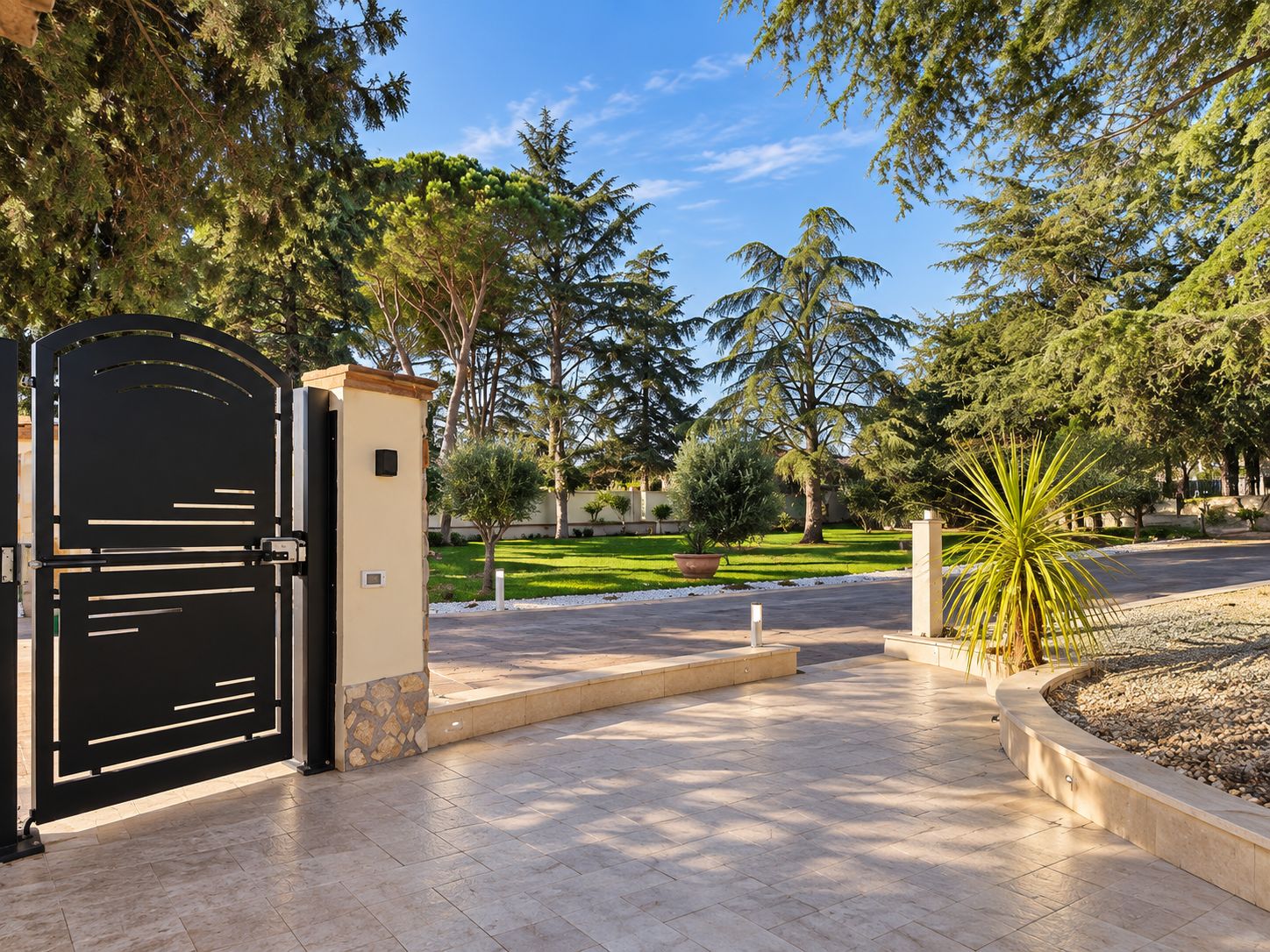 A gated entrance with a paved pathway leading to a landscaped area with trees and a palm plant.