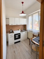Cocina pequeña con gabinetes blancos, estufa, lavamanos y ventana con cortinas de encaje.