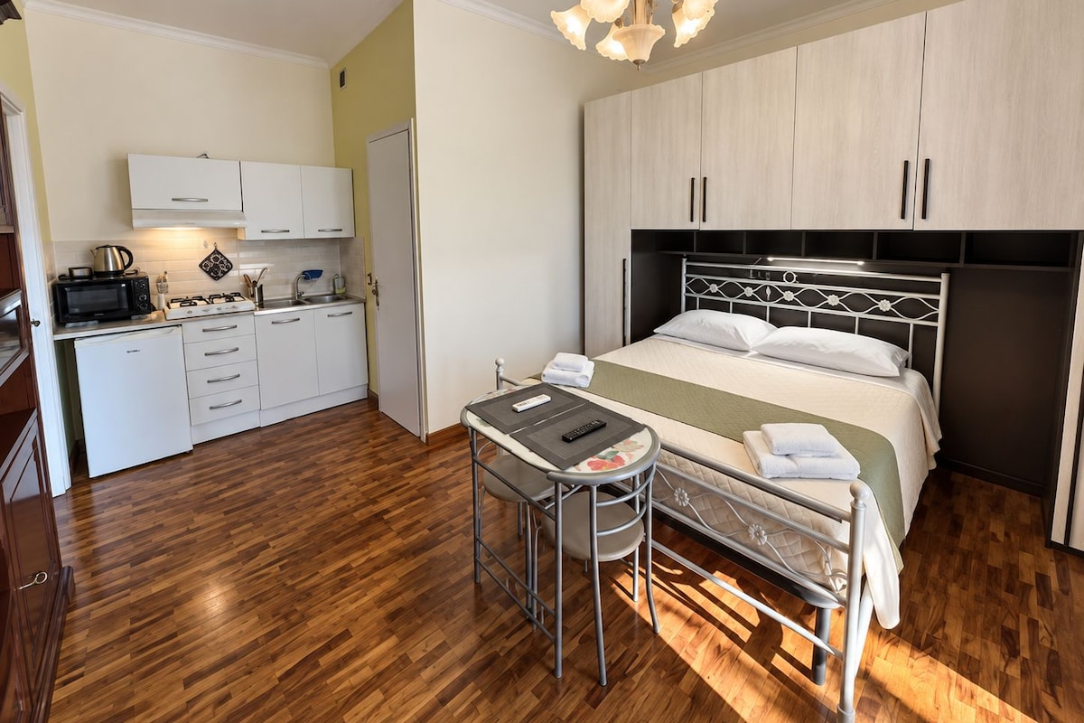 Une chambre d’hôtel compacte comprenant un coin cuisine, un lit avec une tête de lit, une petite table avec une chaise et un luminaire au plafond.