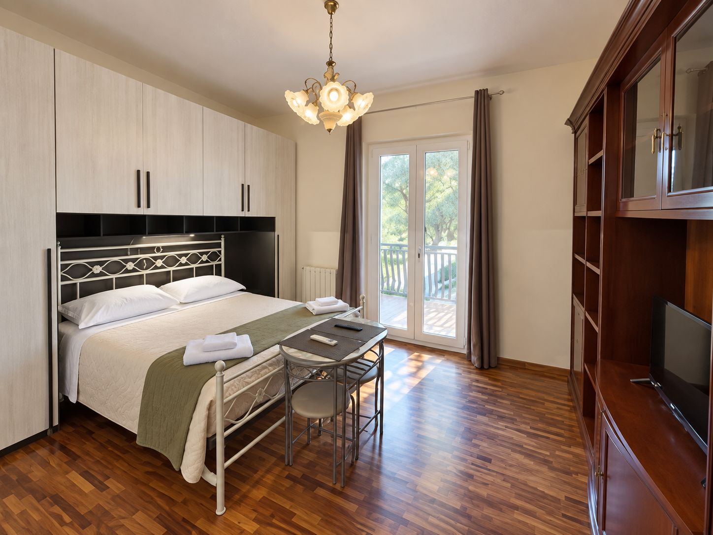 A hotel room with a bed, a desk, a chair, a television, and a balcony with curtains.