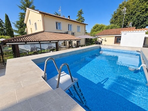 Une piscine avec un escalier, entourée d’une terrasse couverte et d’une maison dont le toit est recouvert de tuiles.