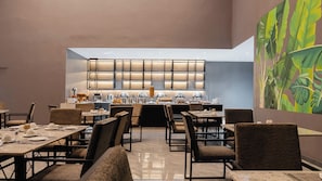 A modern dining area with multiple tables and chairs, a buffet counter with various food items, and a large window with a grid pattern. - The Urban Stay Kalumbila (Kalumbila)