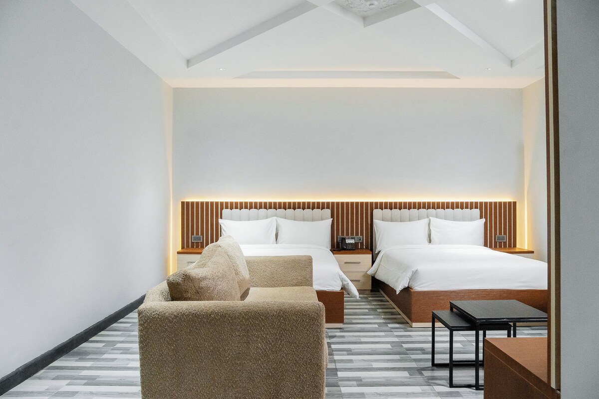 A modern hotel room with two beds, a sofa, a small table, and a nightstand.