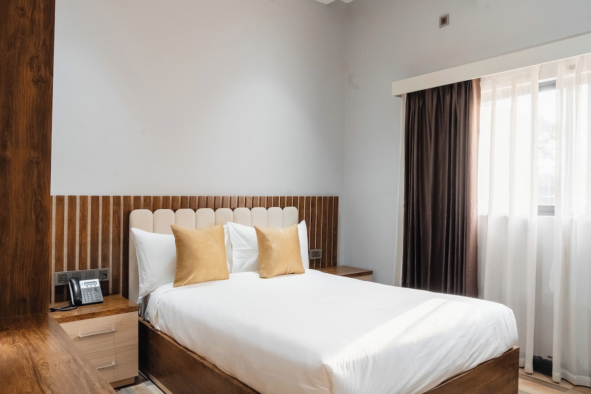 A hotel room with a wooden headboard, a bed with white bedding and two beige pillows, a bedside table with a telephone, and a window with curtains.