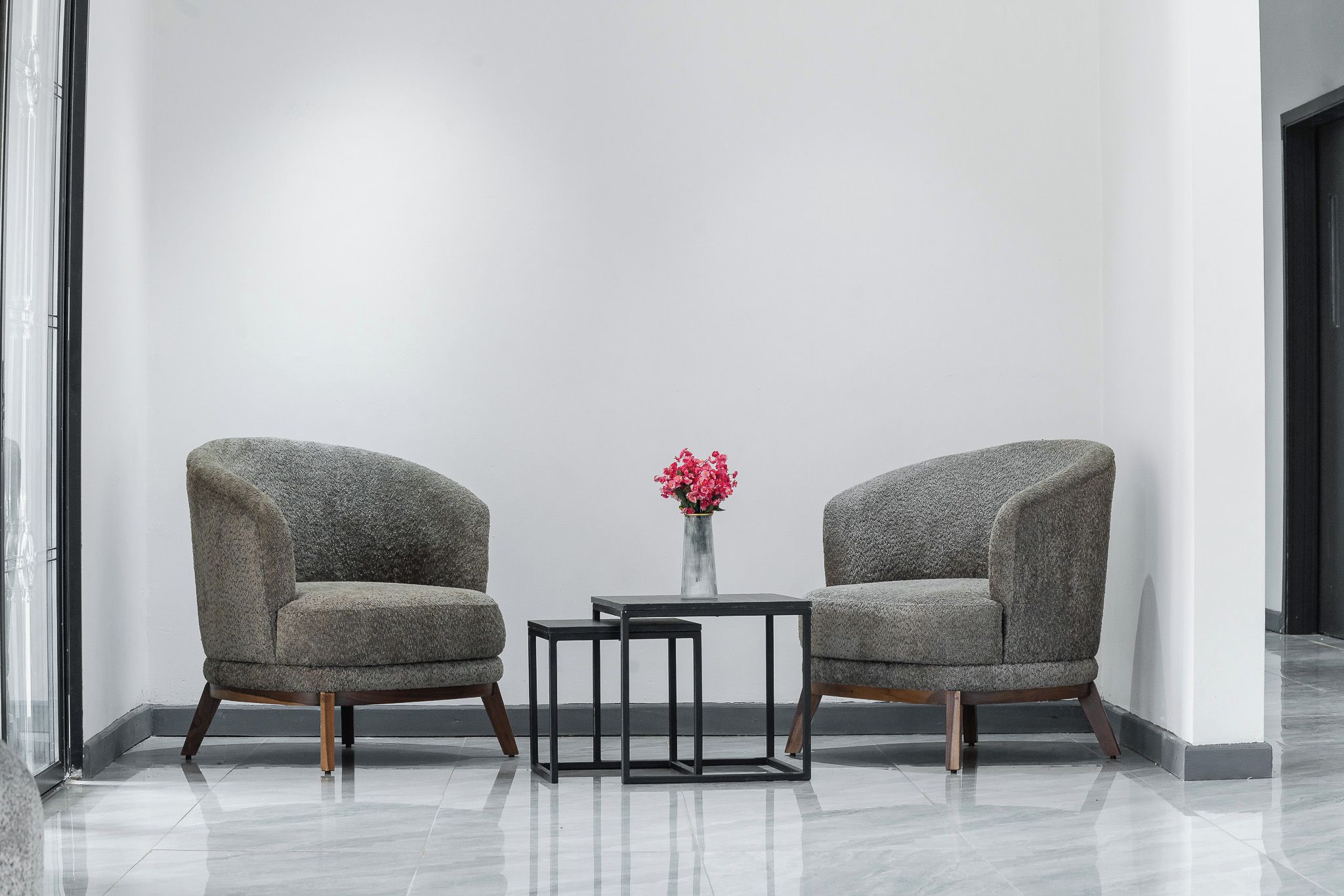 Two armchairs with wooden legs, a small table, and a vase with pink flowers in a modern room with a plain white wall and a glass door.