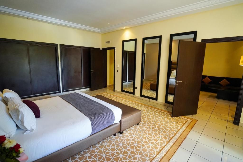 Una habitación de hotel con cama, espejo grande, armario y zona de estar con cojines.