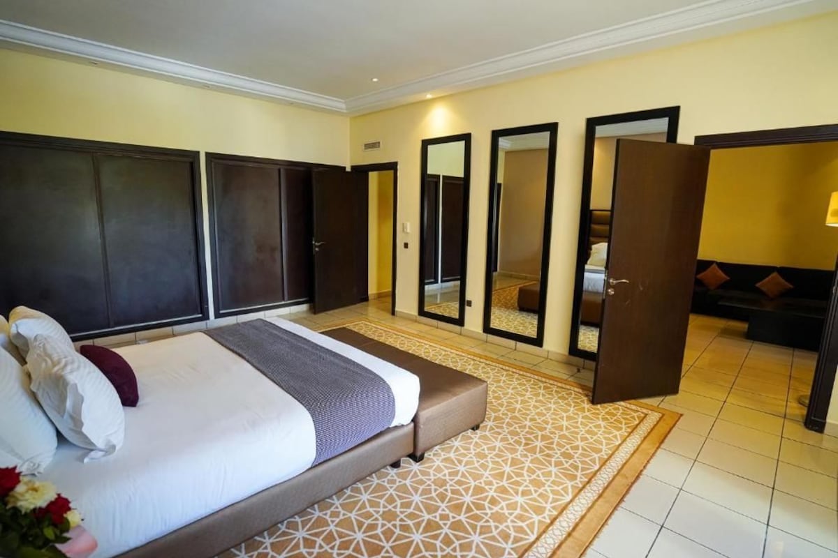 Una habitación de hotel con cama, espejo grande, armario y zona de estar con cojines.