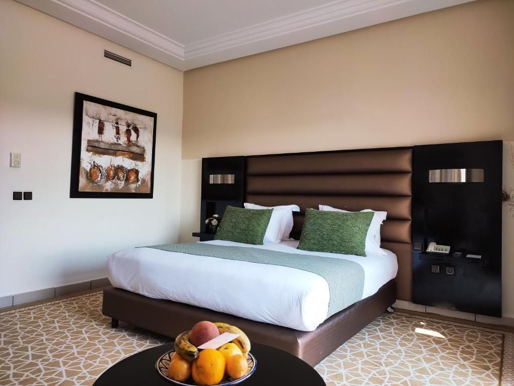 Une chambre d’hôtel avec un grand lit, une tête de lit dotée de commandes intégrées, une œuvre d’art encadrée au mur et un panier de fruits posé sur le sol.