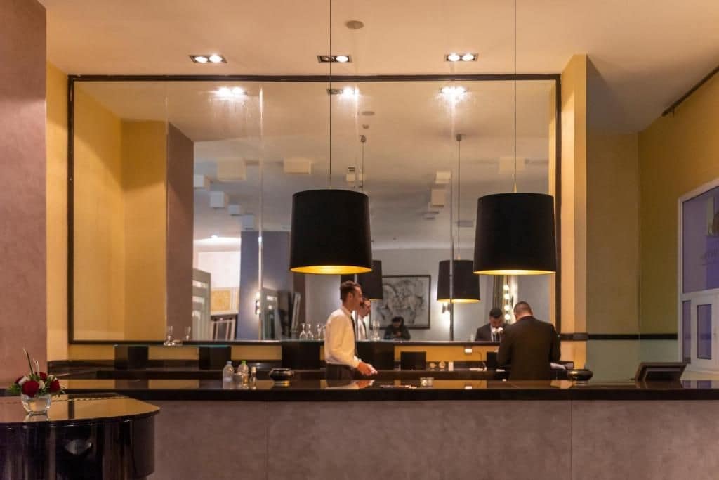 Zona de recepción de hotel con un espejo grande, dos lámparas colgantes negras, un mostrador de recepción y una mesa redonda con un florero.