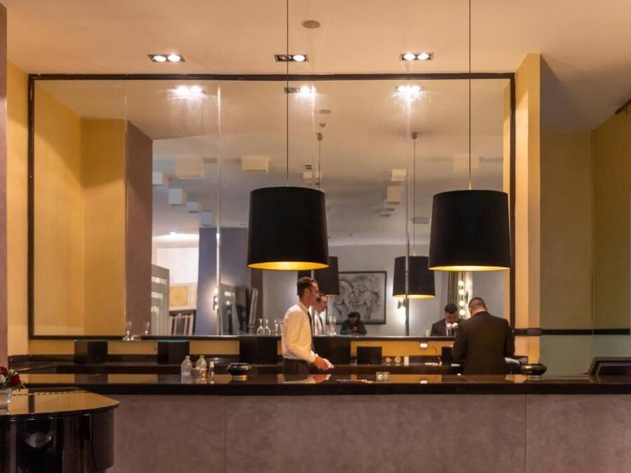 A hotel reception area with a large mirror, two black pendant lights, a reception desk, and a round table with a vase of flowers.