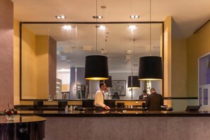 Zona de recepción de hotel con un espejo grande, dos lámparas colgantes negras, un mostrador de recepción y una mesa redonda con un florero.
