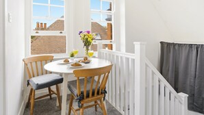 A small dining area with a round table, two wooden chairs, a vase of flowers, and a window with a view of brick buildings. - Heritage Room - (Egham Station 1 min walk) (Egham)