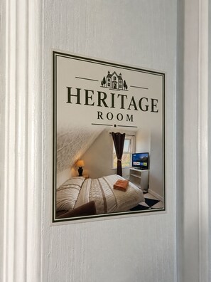 A framed sign with the text 'Heritage Room' and a depiction of a bedroom with a bed, a TV, and a window.