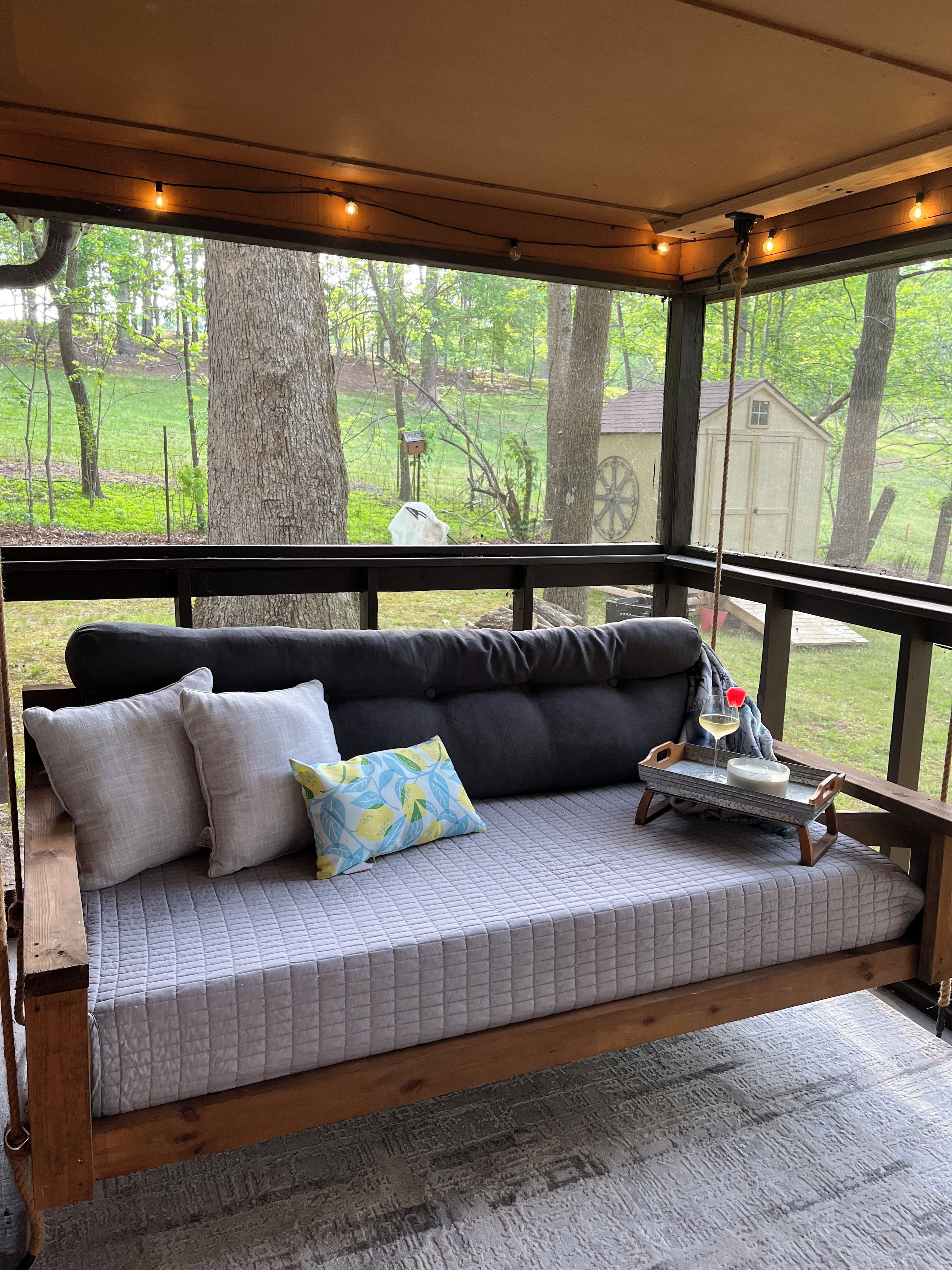 A wooden deck with a cushioned bench, a tray with a candle and a glass, and a view of a green landscape.