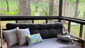 A wooden deck with a cushioned bench, a tray with a candle and a glass, and a view of a green landscape.