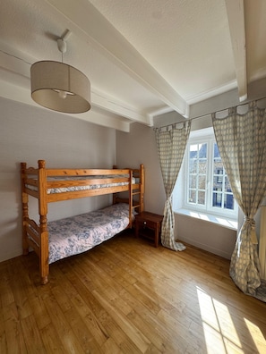 Una habitación con literas, suelo de madera, ventilador de techo, ventana con cortinas y una pequeña mesa.