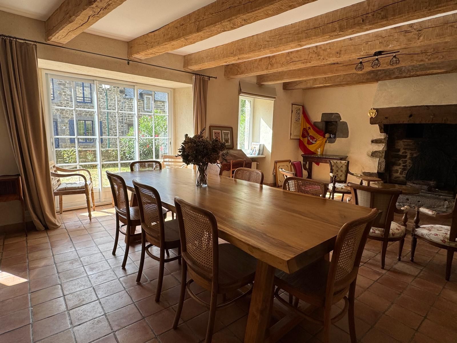 Un espace repas comprenant une grande table et des chaises en bois, une cheminée en pierre et de grandes fenêtres avec des rideaux.