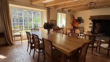 Un espace repas comprenant une grande table et des chaises en bois, une cheminée en pierre et de grandes fenêtres avec des rideaux.