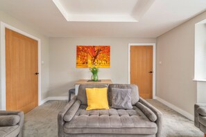 A neutral-toned living room with a grey sofa, a wooden coffee table, and a painting of autumn trees. - Mill House, Staffordshire (Stafford)