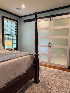 A bedroom with a four-poster bed, a large window with blinds, a nightstand with a lamp, and a patterned rug.