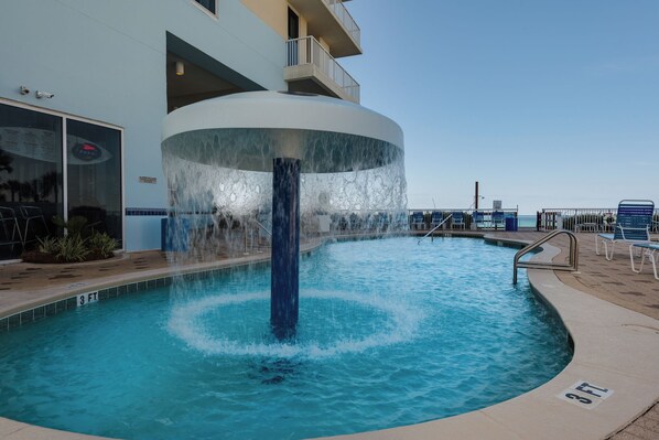 A pool with a central water feature, surrounded by lounge chairs and a building with a balcony. - 17th Floor Gulf Escape | Sleeps 6 | Pools, Hot Tubs & More (Panama City Beach)