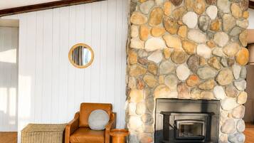 A cozy living room with a stone fireplace, wooden beams, a leather sofa, and a wicker ottoman.