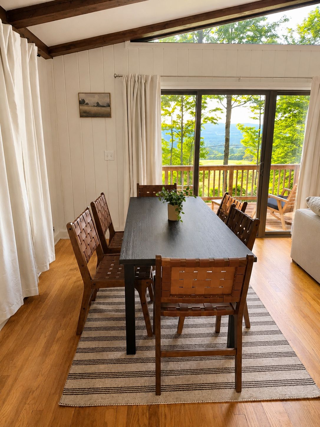 A dining area with a wooden table and chairs, a view of greenery and mountains through large windows, and a balcony with a railing.