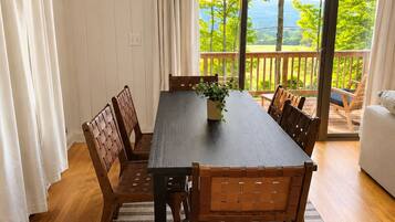 A dining area with a wooden table and chairs, a view of greenery and mountains through large windows, and a balcony with a railing.