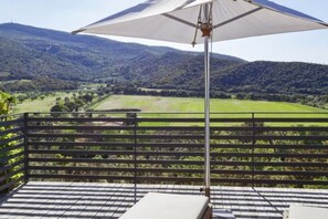 A terrace with a view of a green valley and hills, a metal railing, and a large umbrella.
