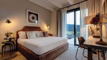 Ein Hotelzimmer mit einem großen Bett, einem Schreibtisch, einem Sessel und Blick ins Freie, sowie einem an der Wand hängenden Kunstwerk.